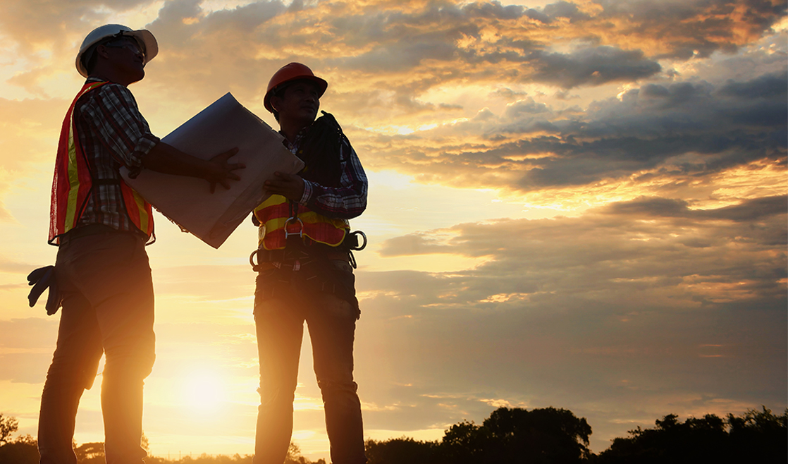 Two construction workers in hard hats and safety vests standing on a construction site at sunset.