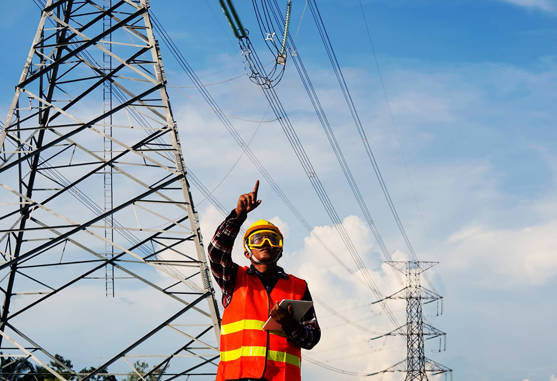 A worker in a hard hat and reflective vest standing in front of a large electrical tower.