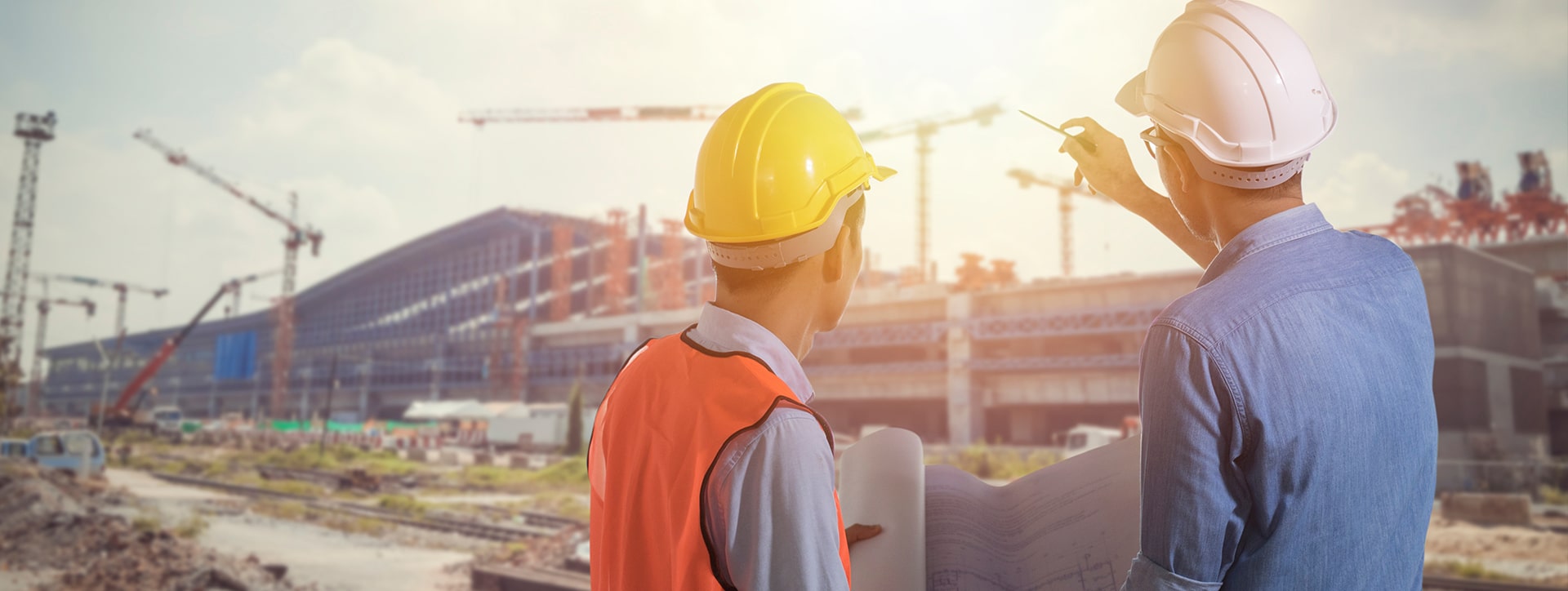 two construction workers looking at a construction site. The worker in the yellow hard hat is holding a rolled up blueprint. The worker in the white hard hat is pointing towards the construction site. 