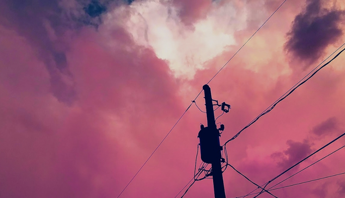 A photo of a telephone pole with power lines against a pink and purple sky. The power lines are black and criss-cross in the sky. 