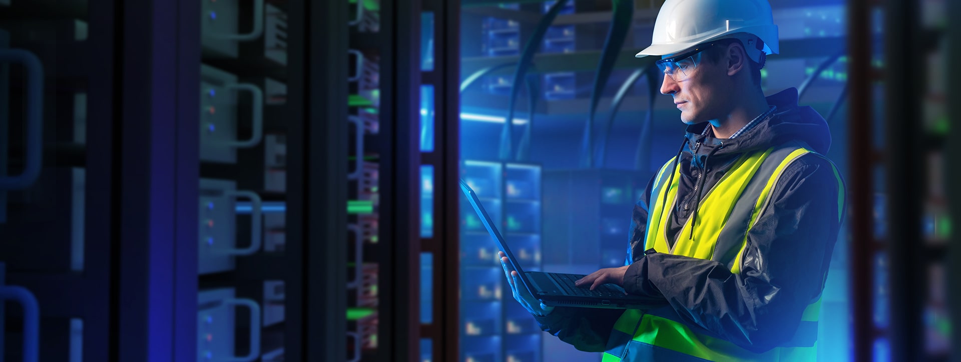 A person in a hard hat and high visibility vest working on a laptop in a server room.